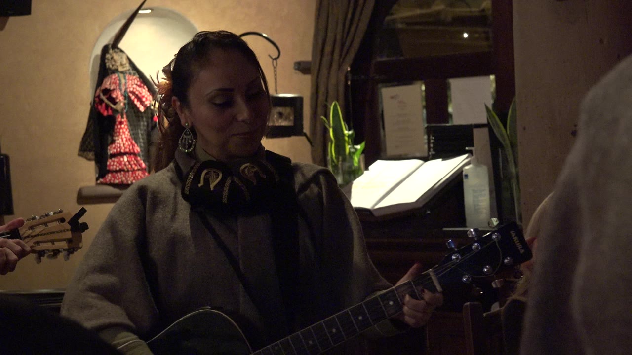 The woman looks down at her guitar while playing indoors in Los Cabos, Mexico, with a pale audience shoulder in the foreground and a music stand or menu behind her.