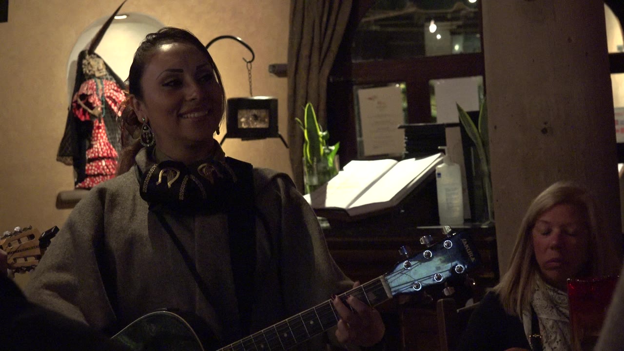 A woman in a gray coat smiles while holding an acoustic guitar during an indoor music moment in Los Cabos, Mexico, with a seated listener and warm background lighting visible.