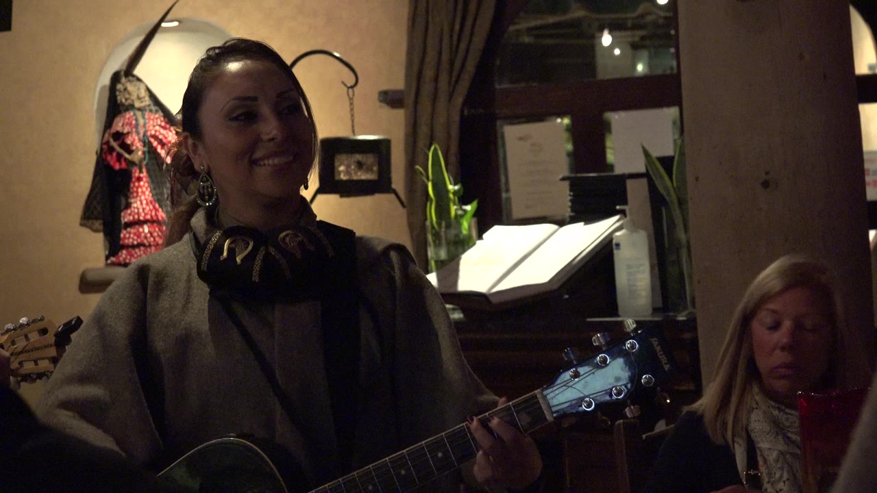 The guitarist remains smiling in the warm indoor Los Cabos setting, with the guitar neck in the foreground and a seated woman behind her near menus and a window.