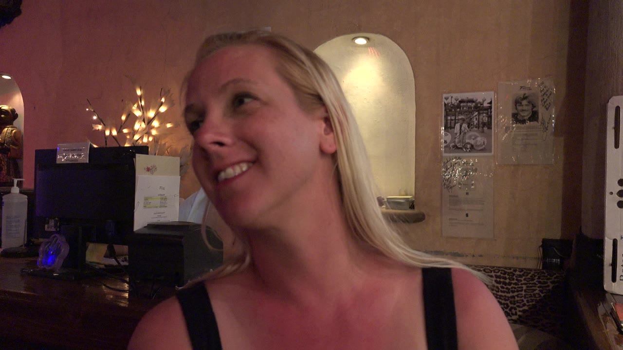 The woman remains close to camera and smiles off to the side in the Los Cabos restaurant, with warm lights glowing behind the counter.