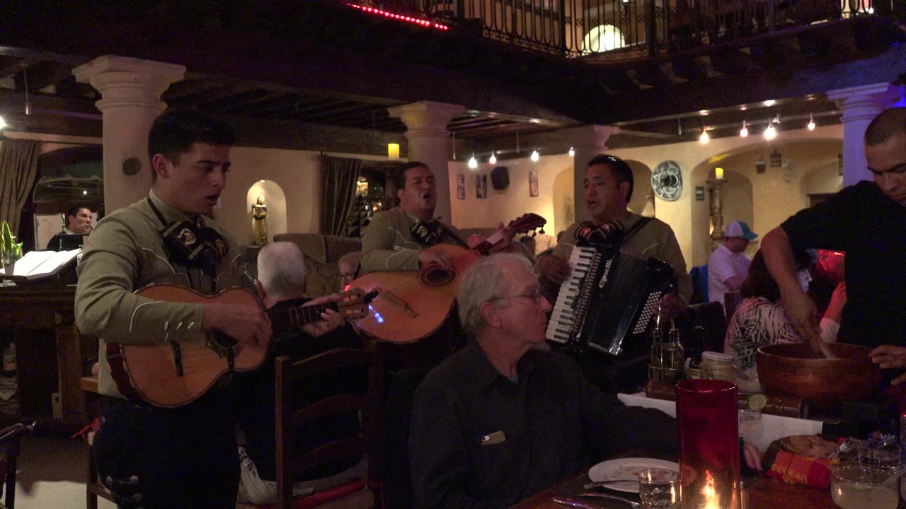 In Los Cabos, Mexico, musicians with guitars and an accordion perform near seated diners at a warmly lit bar table.
