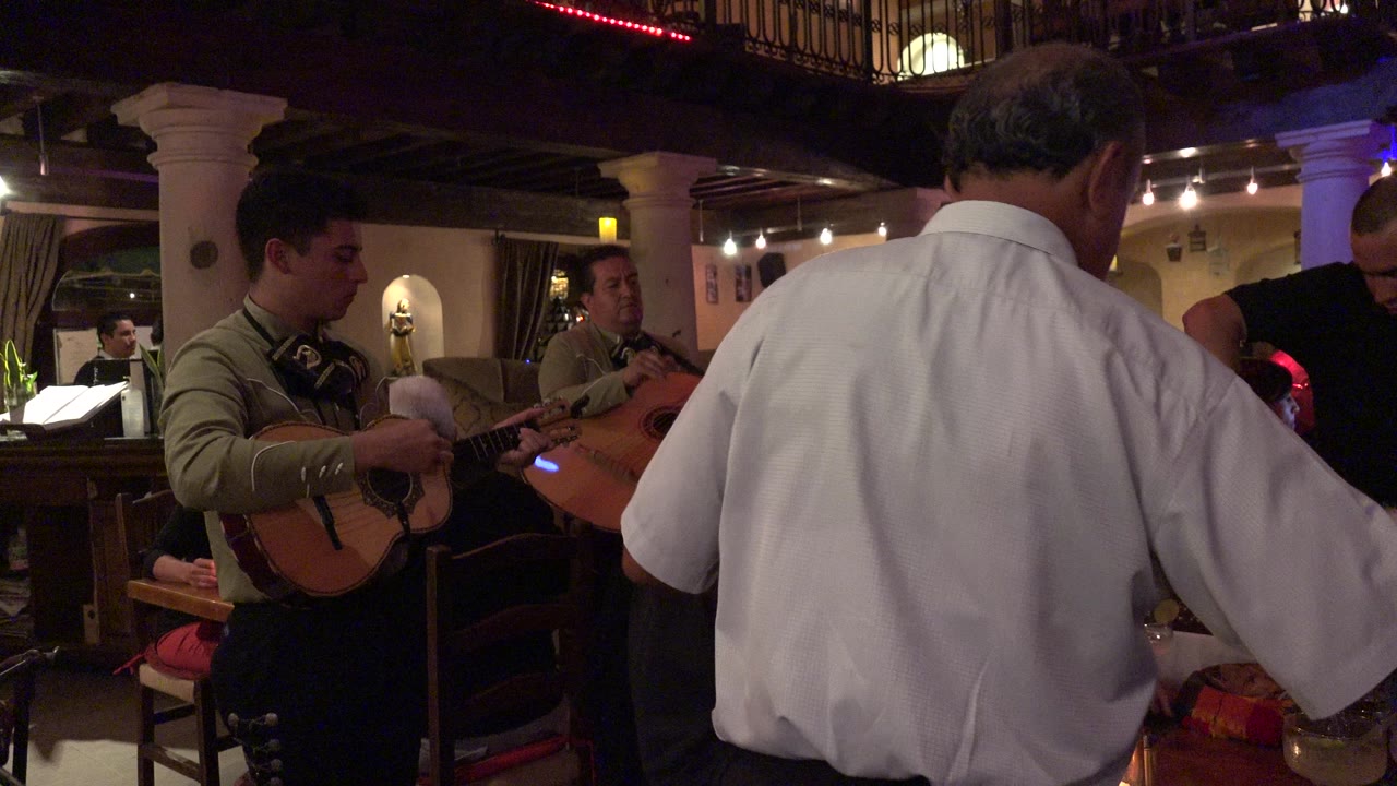 In a Los Cabos restaurant, two musicians with string instruments stand near tables while a man in a white shirt fills the foreground with his back to camera.