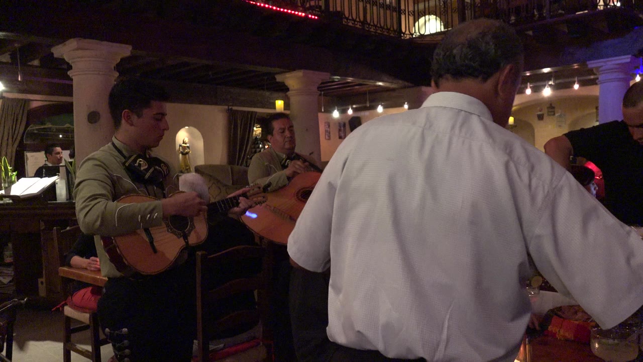 In a Los Cabos dining room, a white-shirted man stands close to camera while musicians with guitars perform beside a table under warm interior lighting.