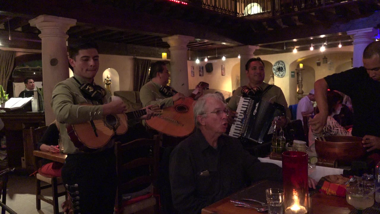 In Los Cabos, Mexico, two musicians with string instruments and a musician with an accordion stand near a seated older man, with warm ceiling lights and dining tables visible.