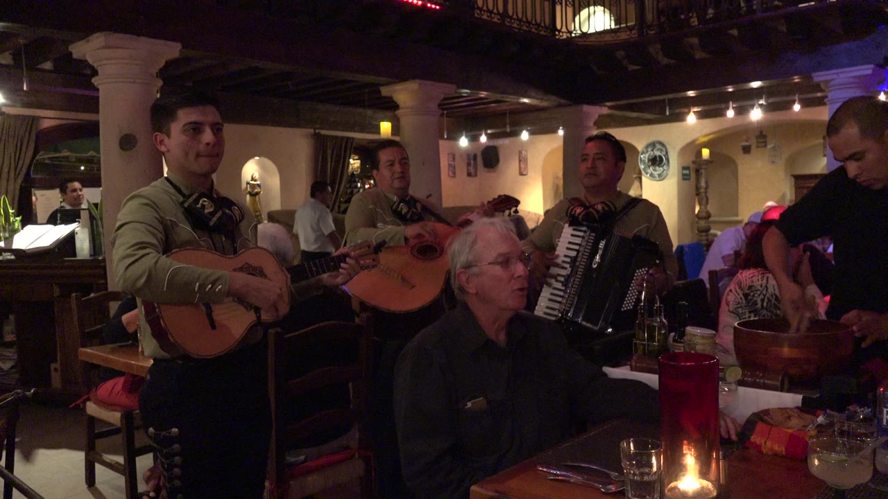 In Los Cabos, Mexico, three musicians stand close to a seated older man at a candlelit restaurant table while diners and warm wall lights fill the background.