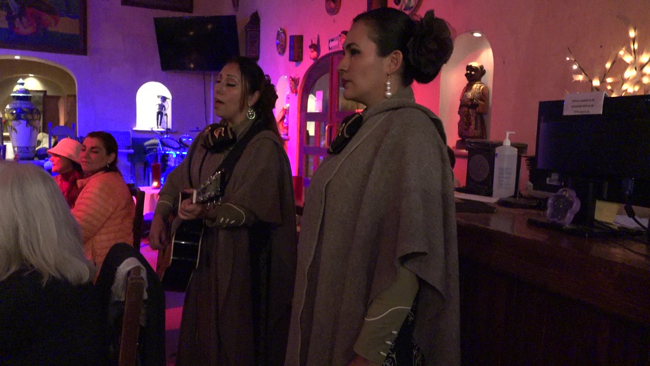 Two women performing beside seated patrons in a colorful pub interior.