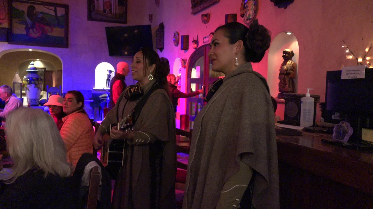 Two women perform inside a Los Cabos club as seated patrons watch under pink and blue interior lighting.
