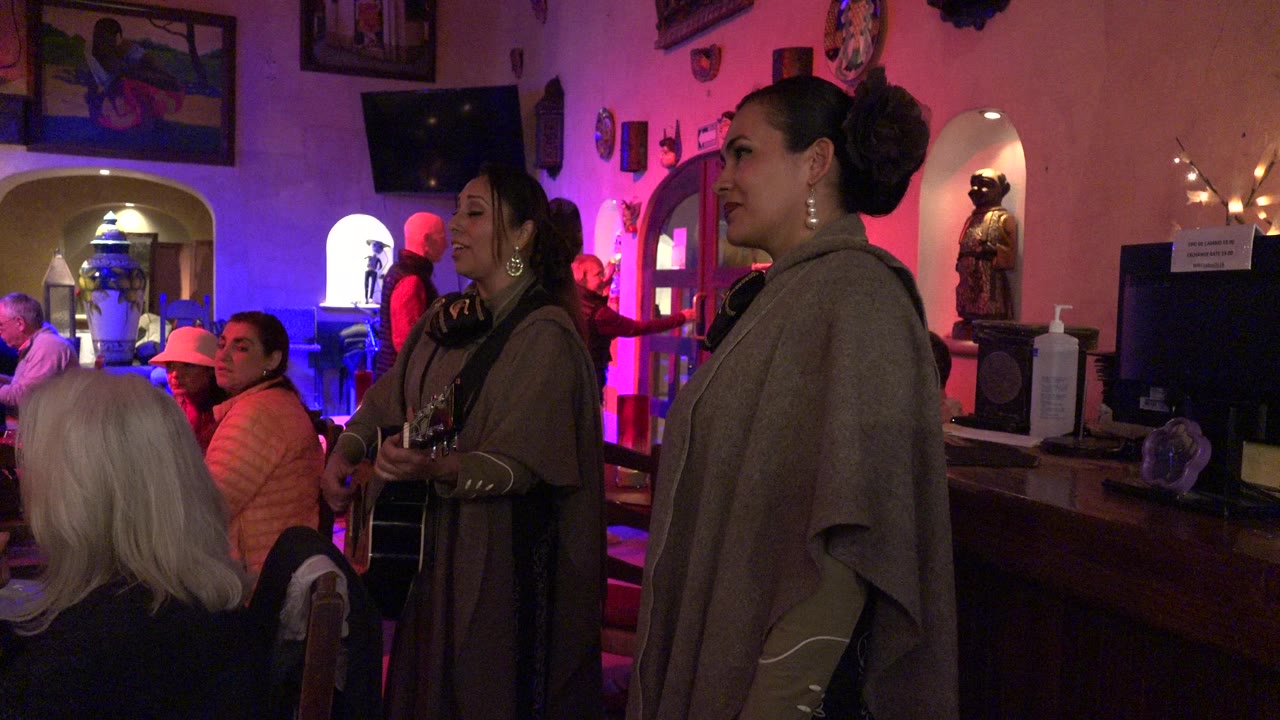 The singer and guitarist remain near the audience in the Los Cabos club, with a red shelf and wall decorations behind them.