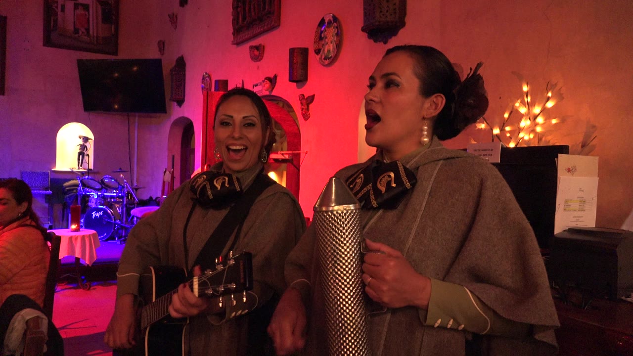 The guitarist smiles toward the camera while the second woman sings beside the metal percussion instrument, with a drum kit and colored lights visible in the background.