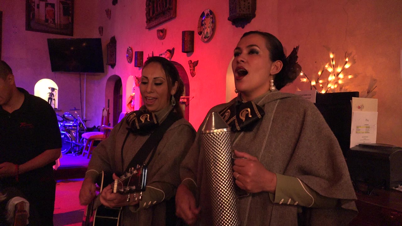Two women sing indoors with guitar and metal percussion.