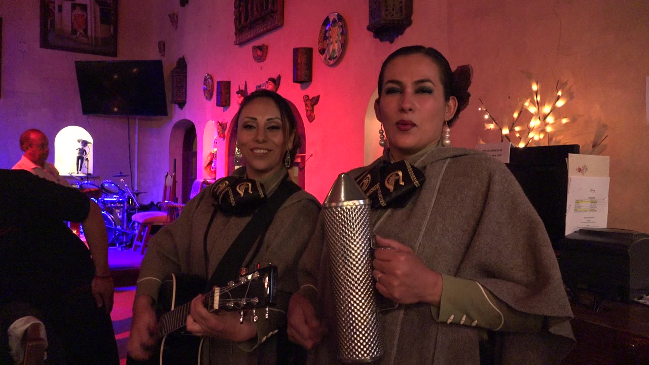 Two women perform with guitar and metal guiro in a colorful indoor room.