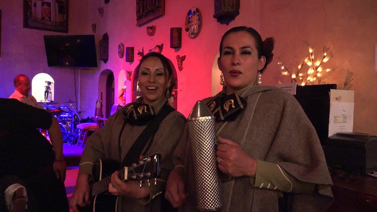 Two women continue singing in the Los Cabos indoor setting, with the guitar angled toward camera and the metal guiro held upright in the foreground.