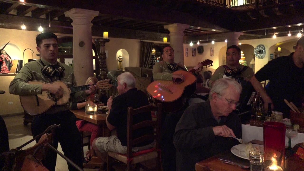 In Los Cabos, Mexico, a guitarist faces forward while two other musicians stand near seated guests and candlelit tables inside the restaurant.