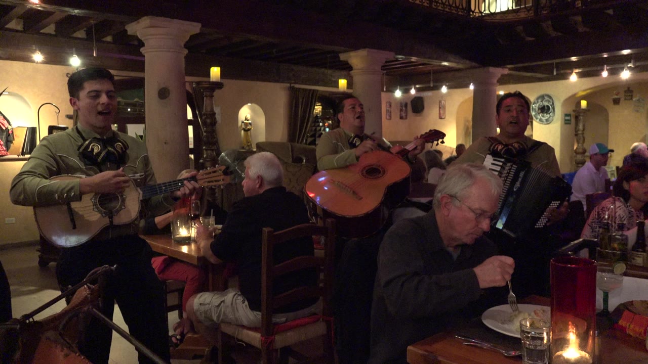 Musicians play beside diners in a warm restaurant interior.