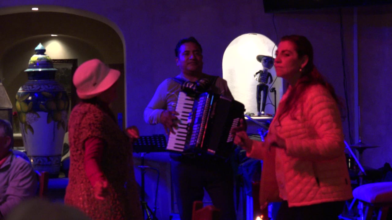 In Los Cabos, Mexico, the woman in a white hat turns toward the accordion player while another woman dances at right, with stage gear and colored lighting visible.