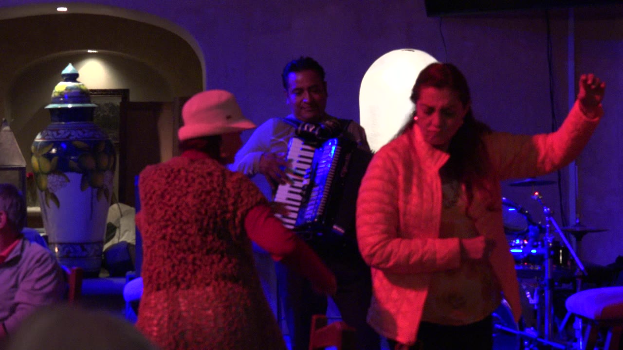 A woman in a white jacket dances in the foreground while another woman in a hat faces the accordion player in the Los Cabos nightclub setting.