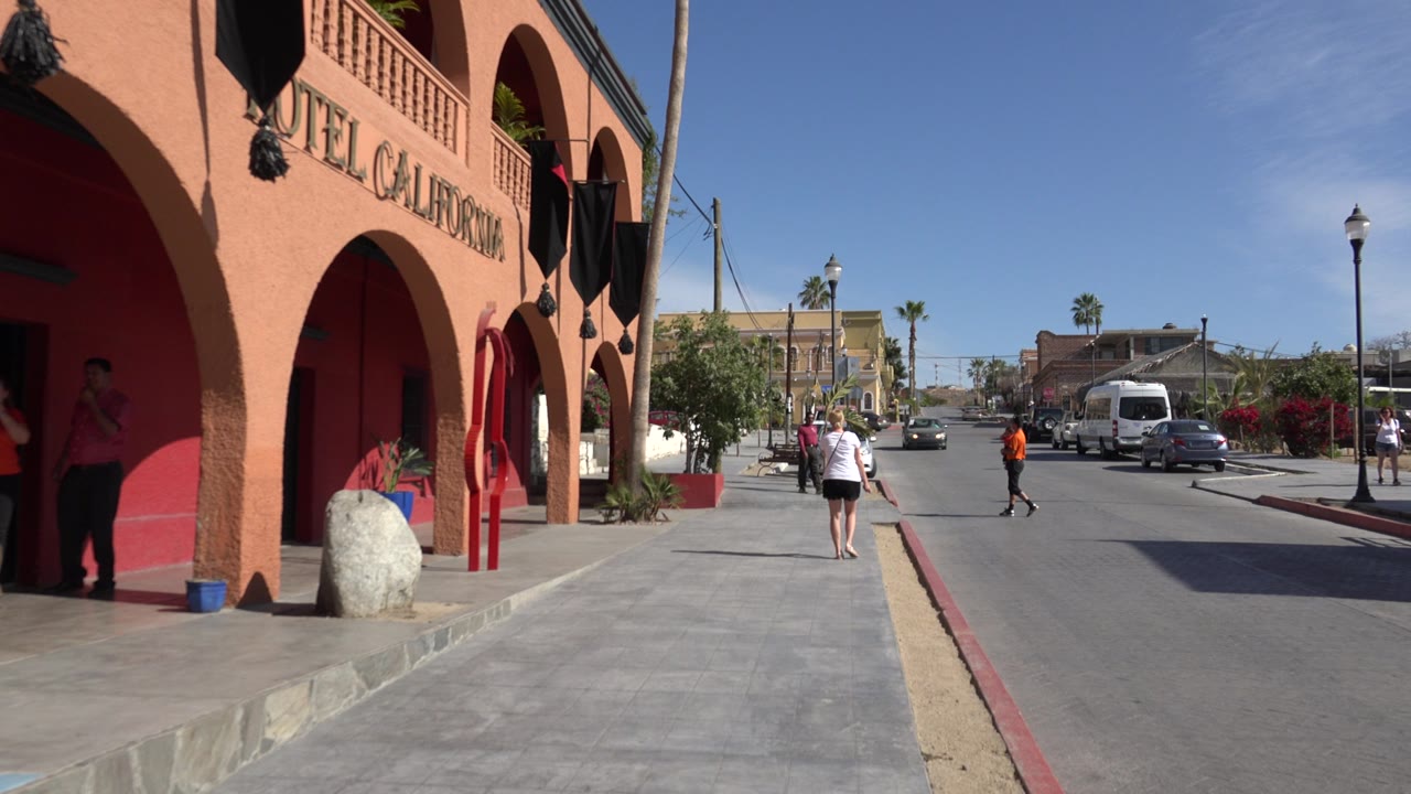 A Los Cabos sidewalk runs past the red and peach facade while an adult in white walks away and vehicles sit along the street.