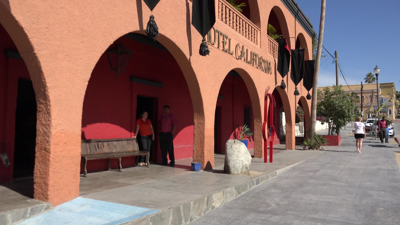 Los Cabos exterior view along orange arches with Hotel California lettering, two people by the entrance, and distant pedestrians on the bright walkway.