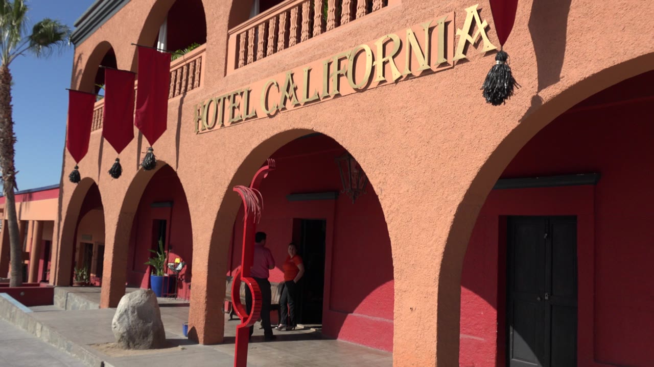 Los Cabos exterior frame centered on the Hotel California facade, with the gold sign across the upper arch and people standing in shadow at the entrance.