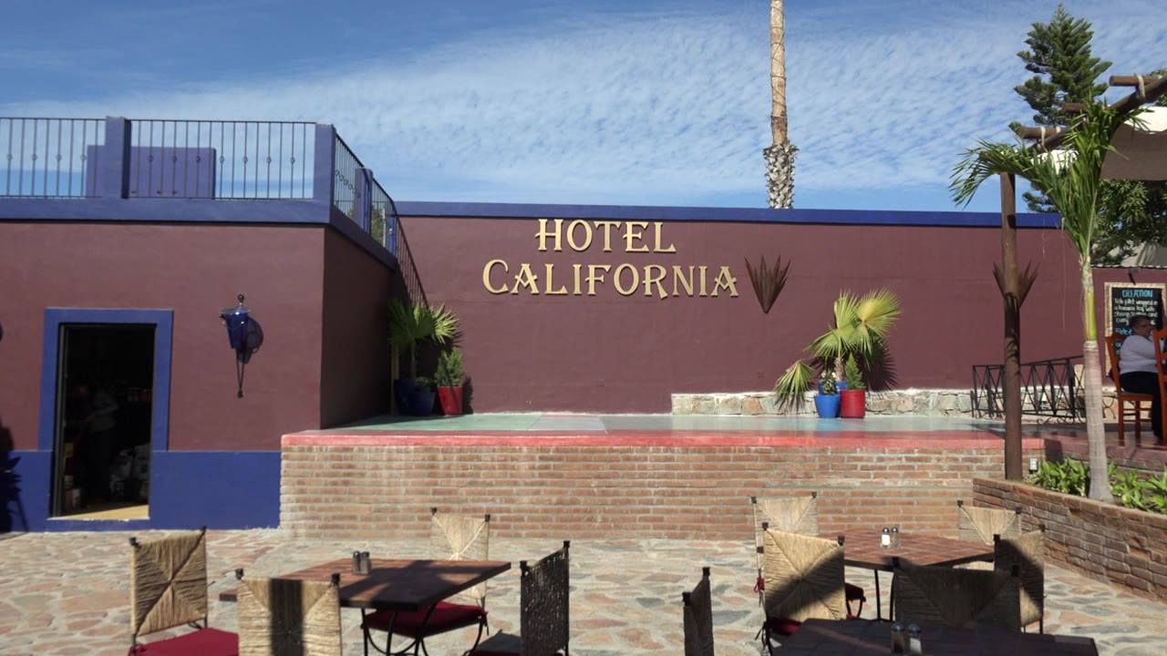 A Los Cabos hotel exterior shows gold Hotel California lettering on a mauve wall, patio seating in the foreground, palms at the right, and people partly visible near the doorway and signboard.