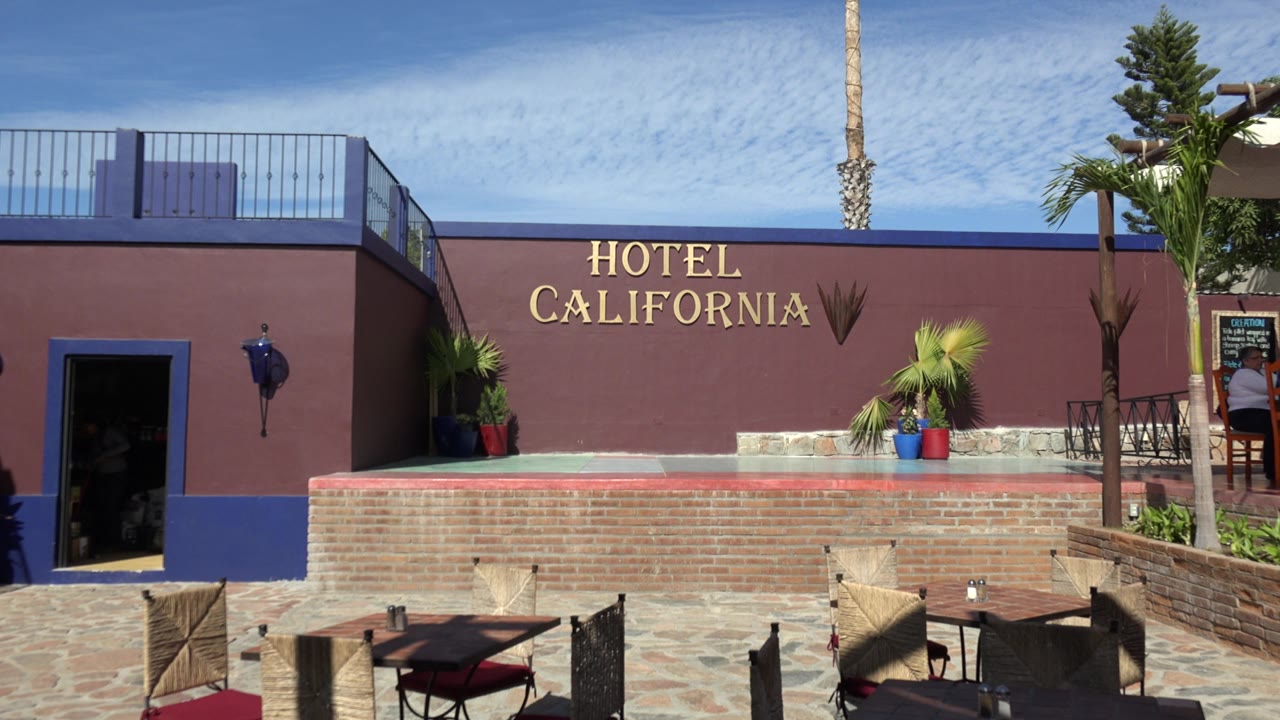 The Los Cabos hotel facade remains centered with the Hotel California lettering above a brick terrace, patio chairs below, and a person visible near the signboard at the right edge.
