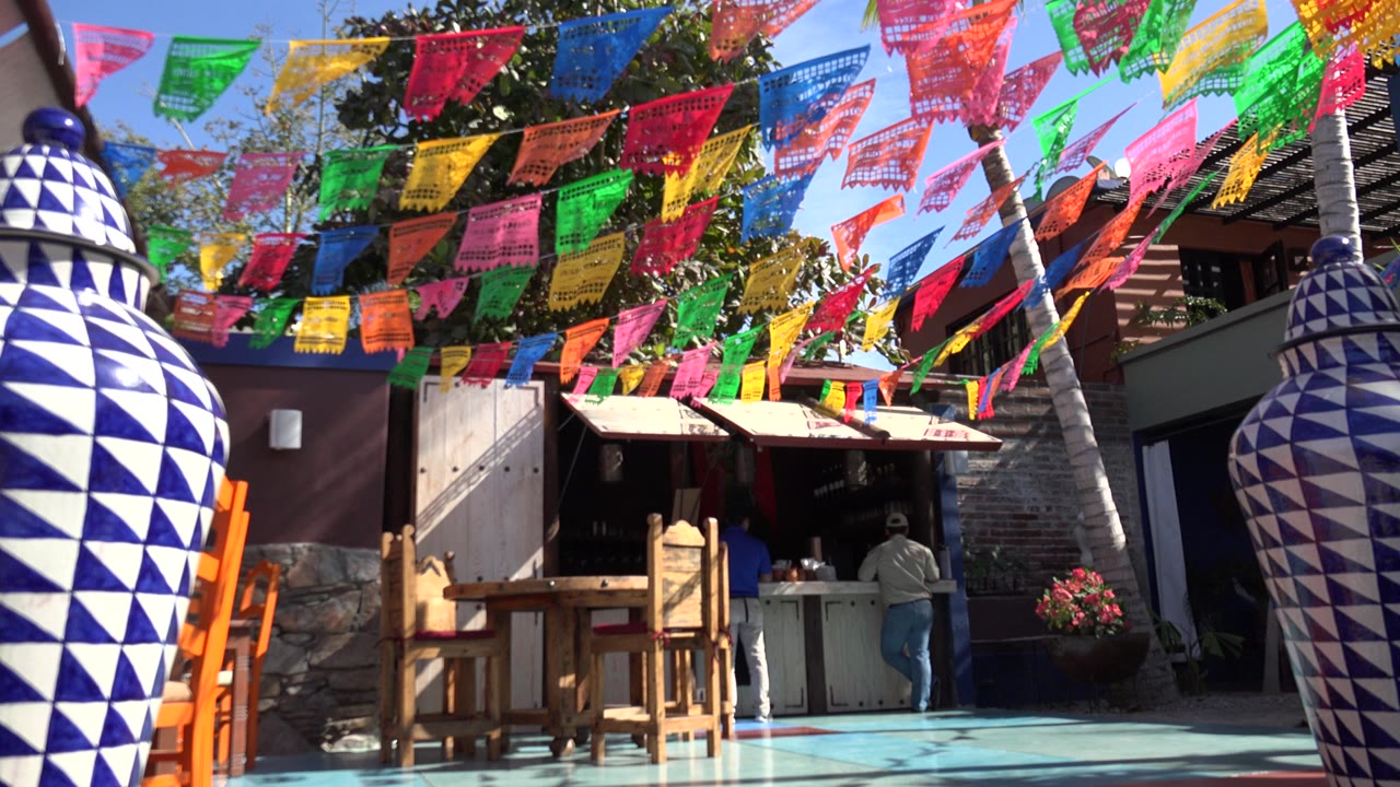 Los Cabos outdoor shop scene with rows of colorful hanging cloths overhead, large blue-and-white patterned pots in the foreground, a wooden table and chairs, and a person partly visible at the counter.
