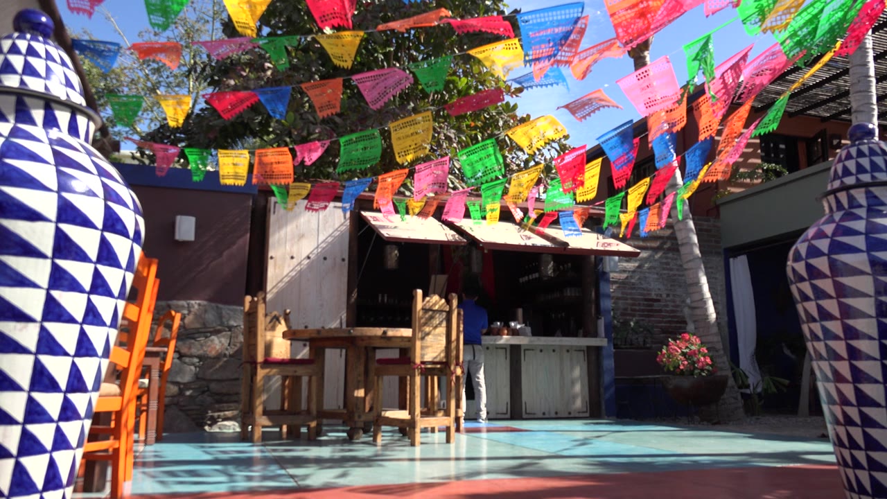 A sunlit Los Cabos courtyard shows rows of bright flags overhead, wooden seating near a shaded service counter, and a large patterned vase at the right edge, with no visible people.