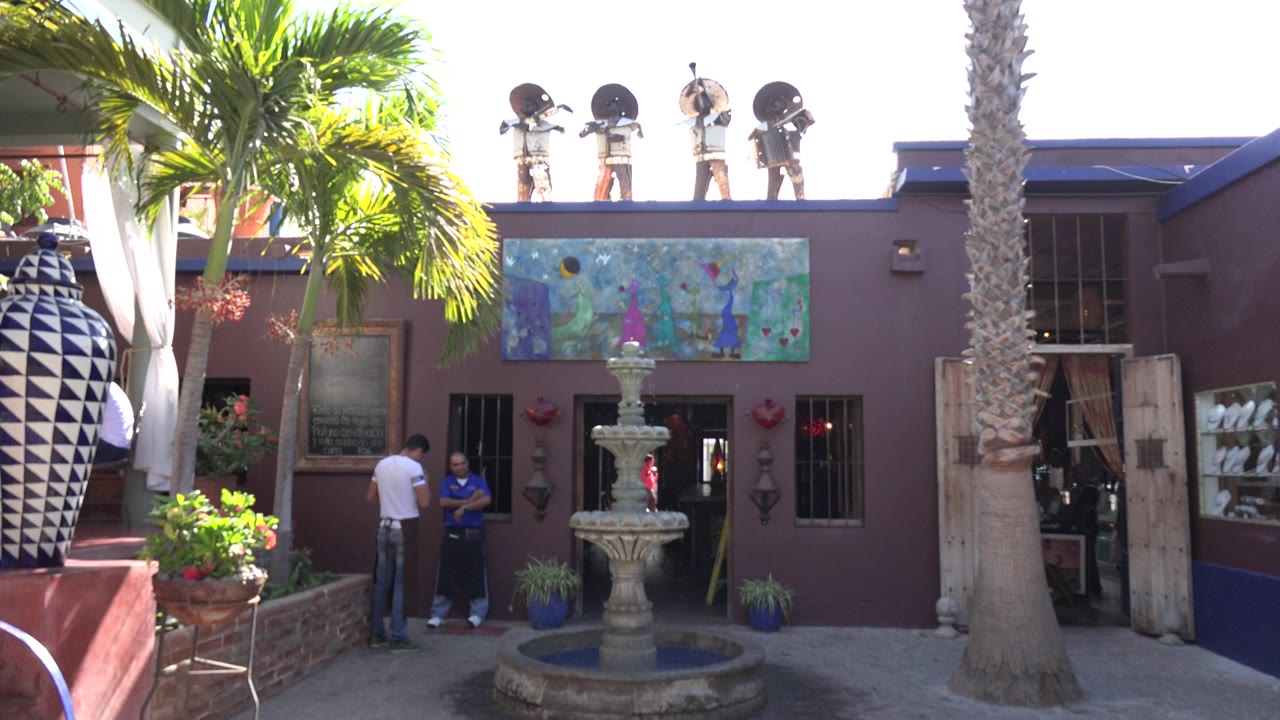 Los Cabos resort courtyard view with the fountain centered, bright palm fronds at left, large black-and-white vases, and two visible people near the entrance.
