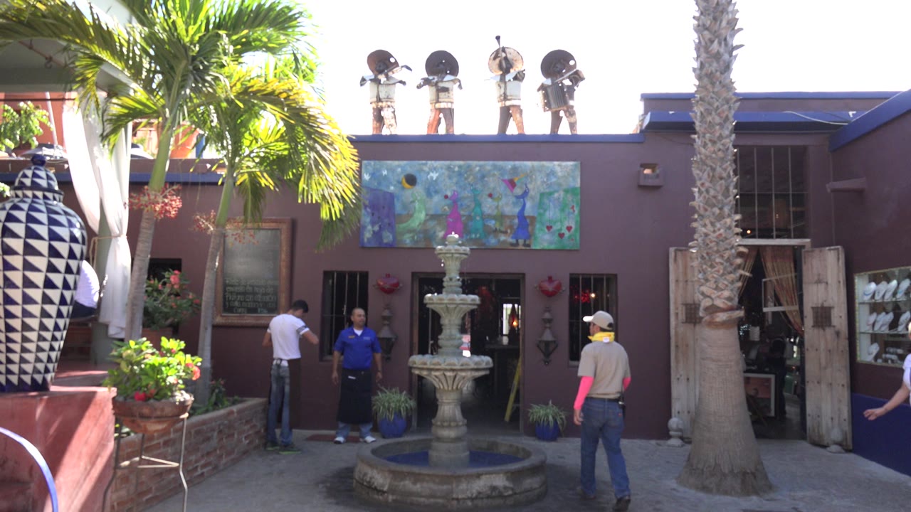 Resort entrance with fountain, palms, rooftop statues, and people near the doorway.