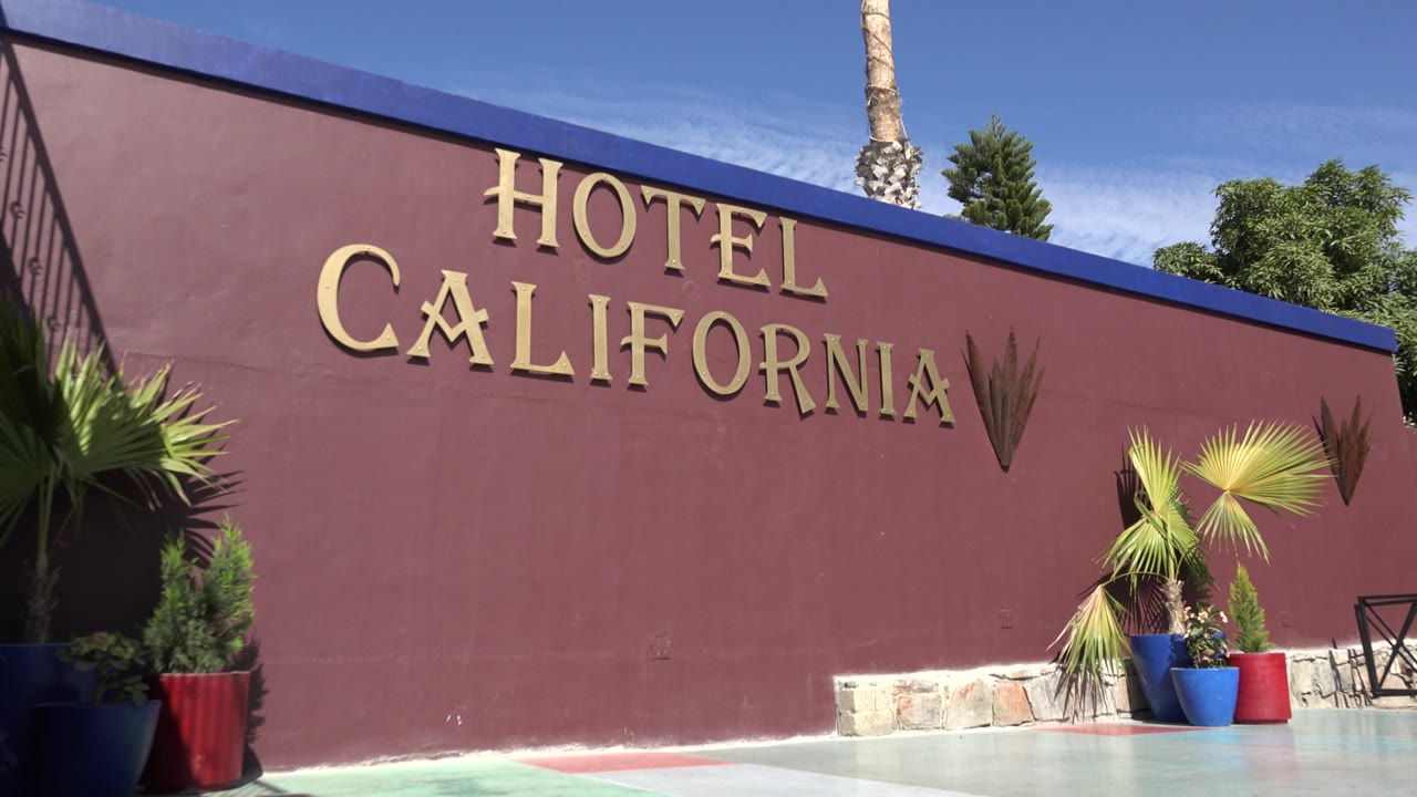 At 0:01, the Hotel California letters remain centered high on the red wall while potted palms and red and blue planters sit near the lower right, with no visible people.