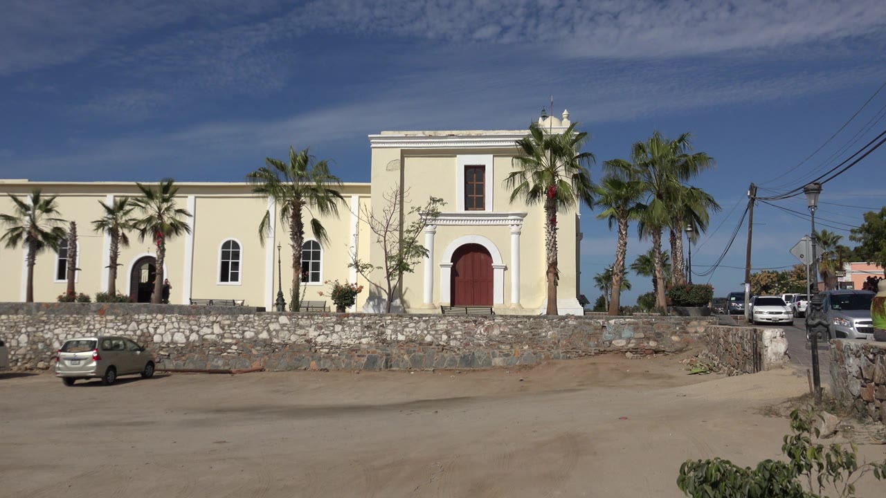 Wide Los Cabos, Mexico view of a pale stucco building with arched dark doorways, a red door, palm trees, a stone wall, parked cars, and no clearly visible pedestrians in the foreground.