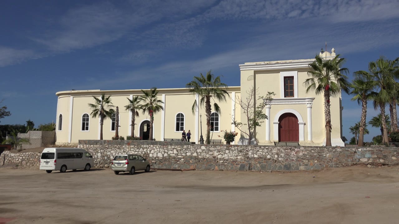 Pale building with red door, palm trees, stone wall, parked cars, and small people near the entrance.