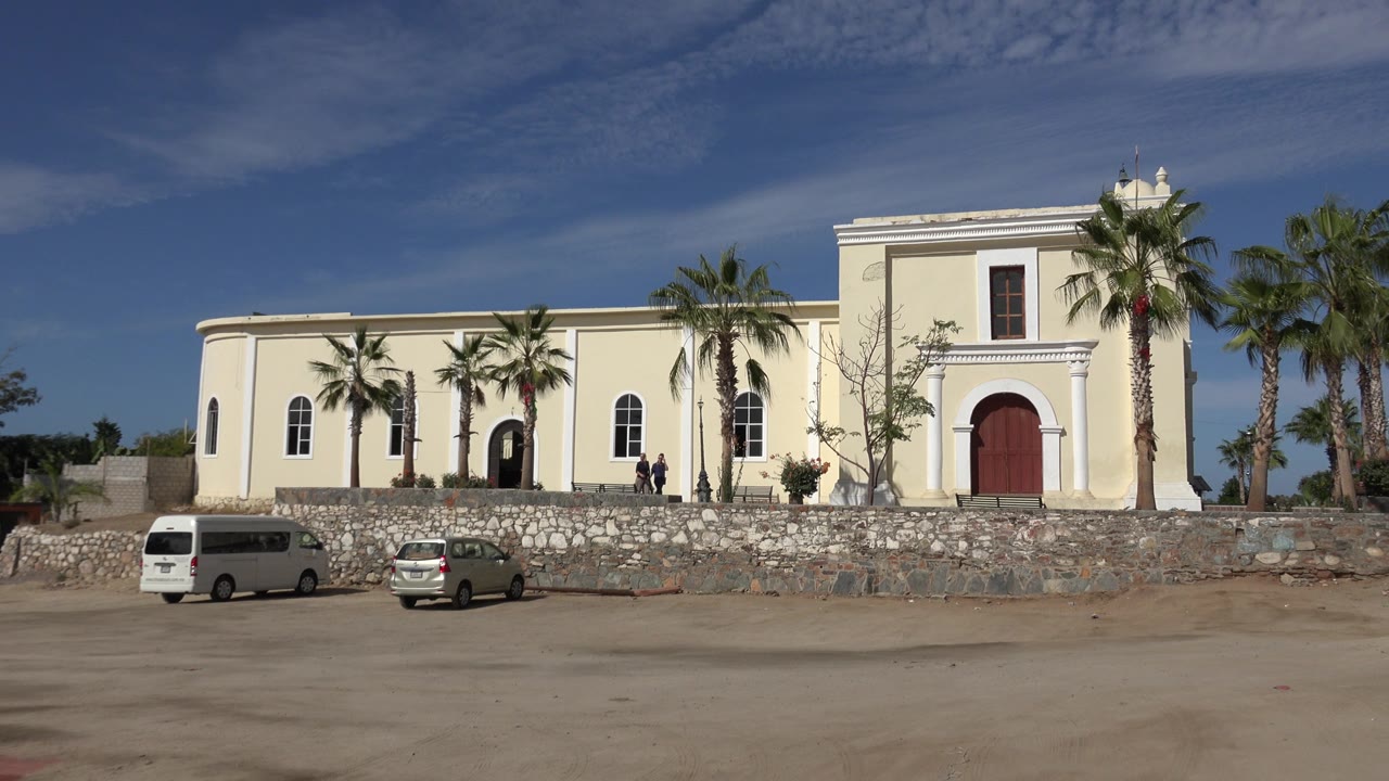 Second Los Cabos frame keeps the building centered, with the red door on the right, palms along the wall, parked white and silver vehicles below, and small visible people by the entrance.