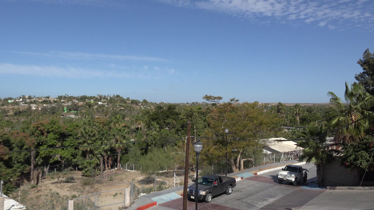 Opening frame of a Los Cabos residential street with a white pickup near a wall, a dark pickup at left, rooftop edges, trees, distant hillside homes, blue sky, and no visible people.
