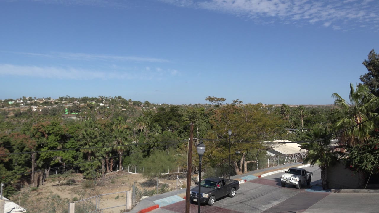 A white pickup has moved farther along the Los Cabos street while the dark pickup remains near the left side, with rooftops, utility poles, dry brush, trees, hills, and no visible people.