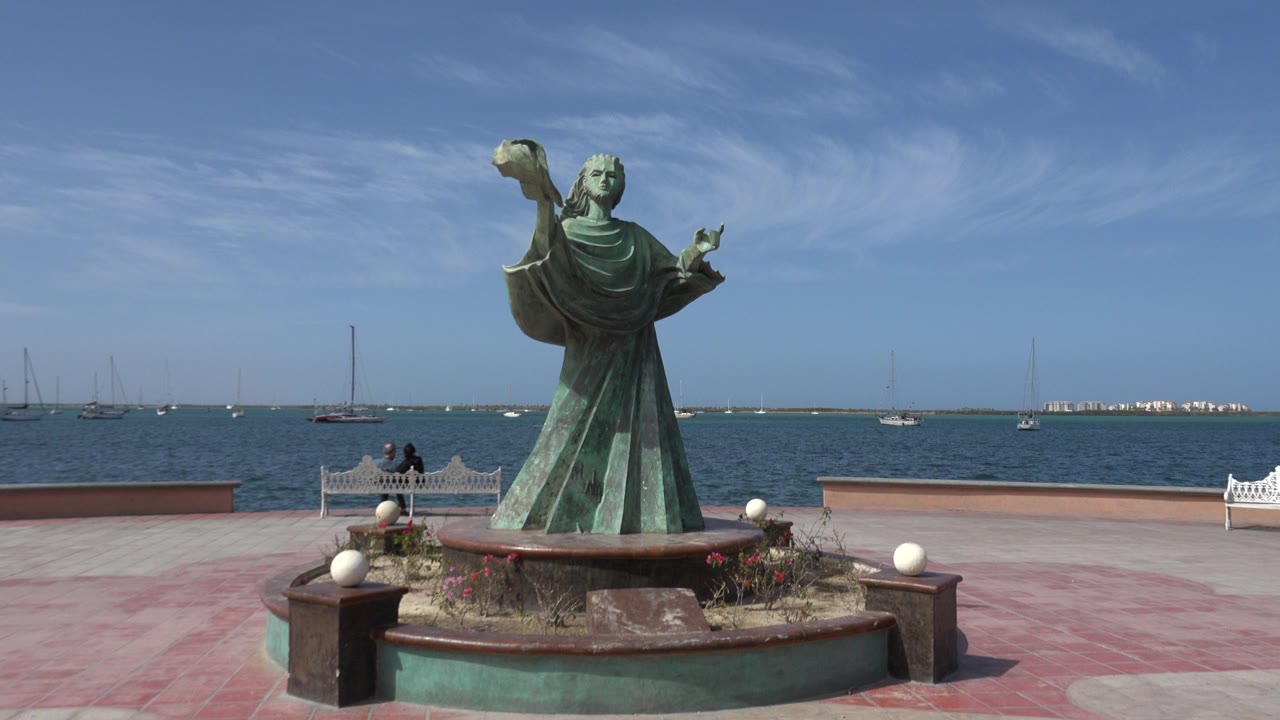 Green copper statue beside a waterfront with boats in the distance.