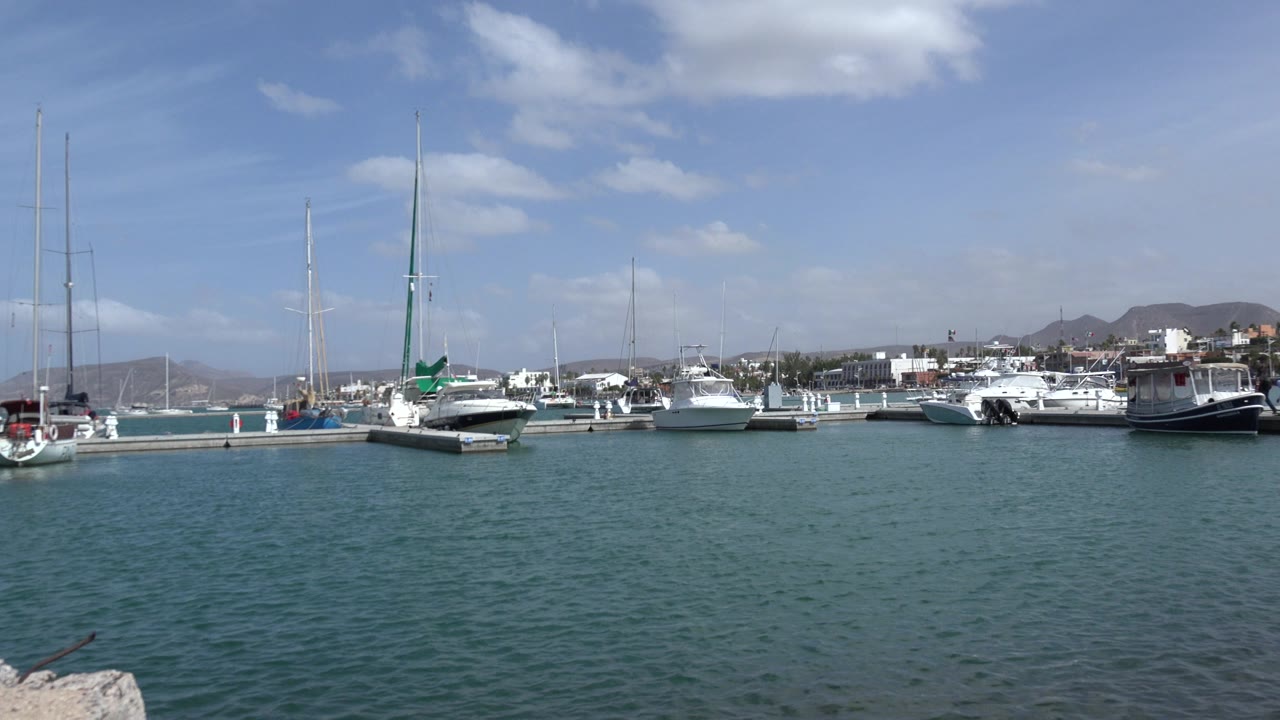 Opening frame of a Los Cabos marina view with sailboats, small motorboats, low hills, blue-green water, scattered clouds, and no visible people.