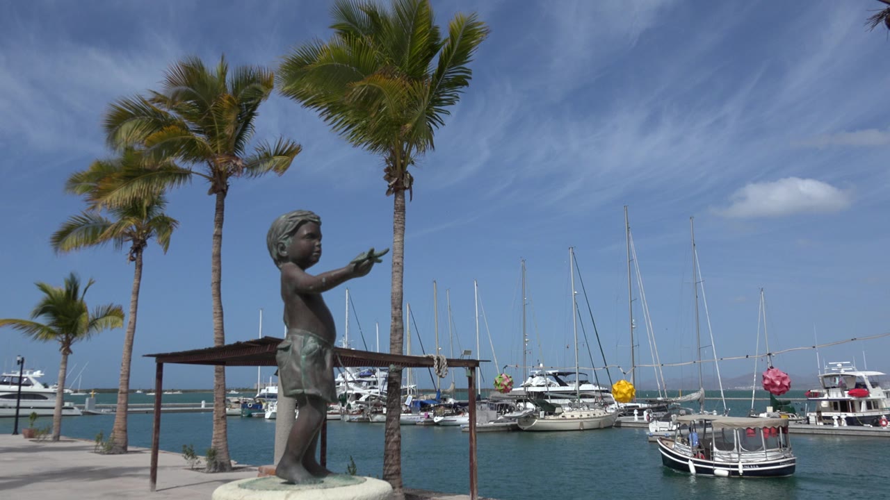Opening frame of a Los Cabos harbor with a bronze child statue in the left foreground, coconut palms above it, anchored boats and sailboat masts across the water, and no live people clearly visible.