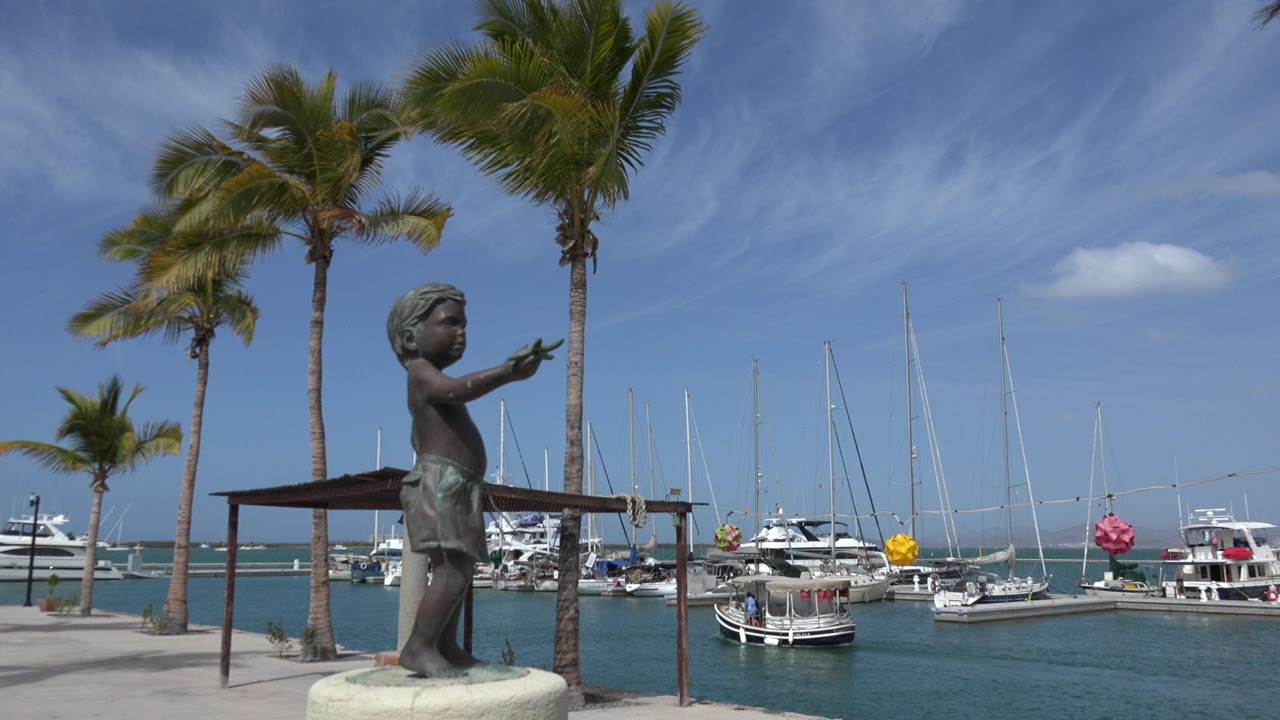 Opening frame of a Los Cabos marina view with a bronze child-like statue in the foreground, palm trees overhead, boats and sailboat masts across blue-green water, and no visible people.