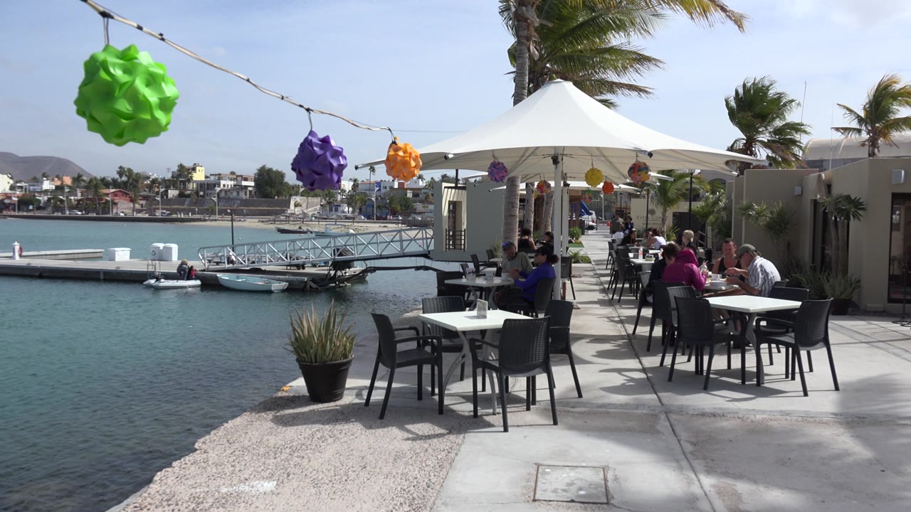 The frame holds on a Los Cabos waterfront restaurant patio, with colorful lanterns crossing the top of the image, diners at tables to the right, and marina water and boats to the left.