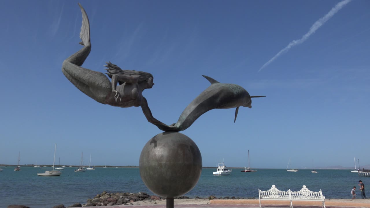 Mermaid and dolphin sculpture beside blue water and anchored boats.