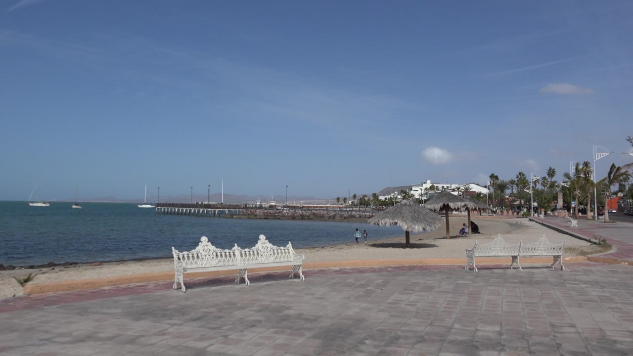 Opening frame of a Los Cabos waterfront promenade with a white bench in front, turquoise water to the left, distant boats, palms, and a few small people near the shoreline.