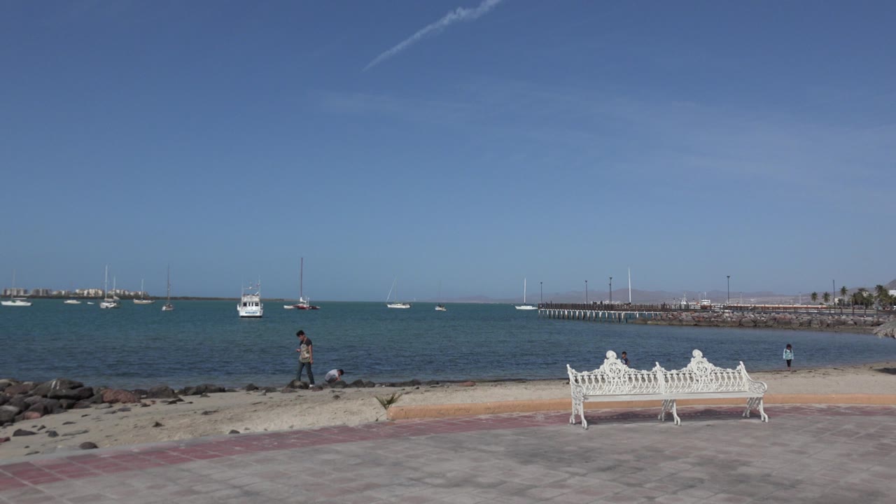 Wide seaside view with white bench, boats, and family near the shore.