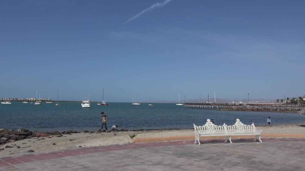 Los Cabos waterfront view with the father standing near the sandy edge, two children lower by the water, and several boats anchored beyond them.