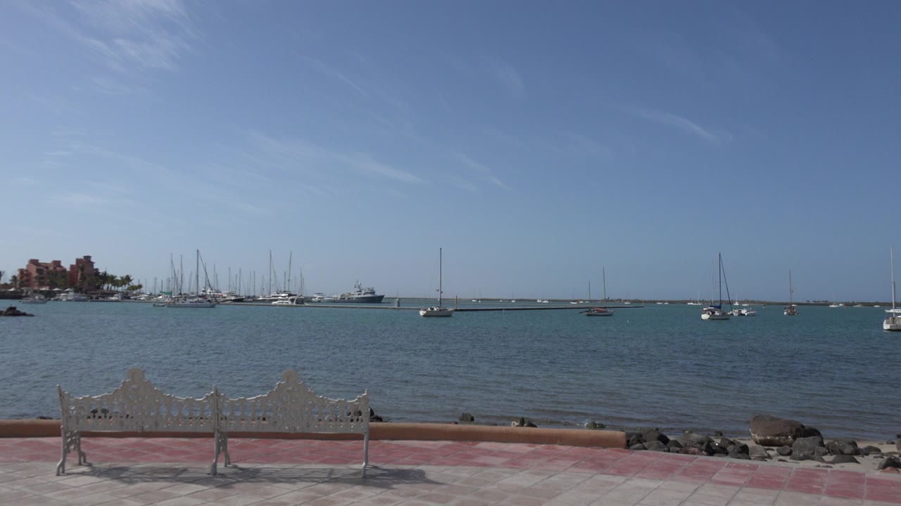 Los Cabos waterfront frame with the bench still centered low in the foreground, small waves on the harbor, sailboat masts, and no visible people.