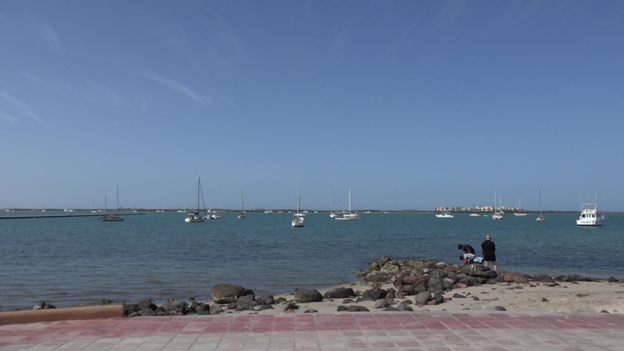 A Los Cabos beach view shows red paving, sand, a rocky ledge, anchored boats on blue water, and two people sitting or standing near the rocks at the start of the clip.