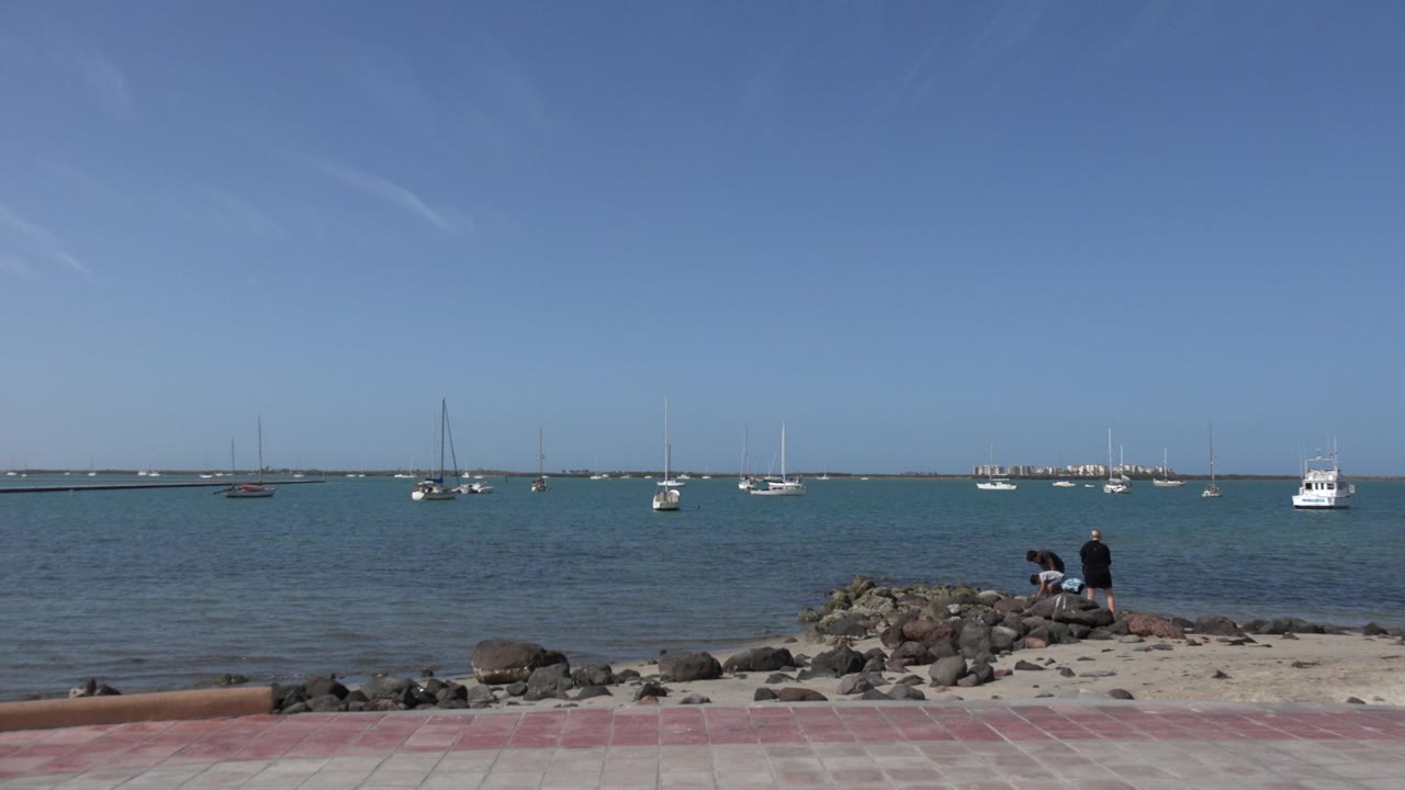 A wide Los Cabos shoreline frame holds the rock ledge in the center, sailboats and small boats across the water, and two people near the rocks under clear blue sky.
