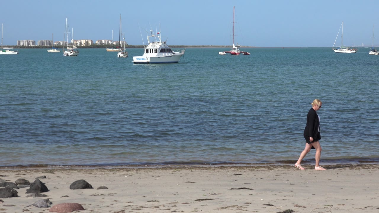 Woman walking barefoot on a sandy beach with boats offshore.