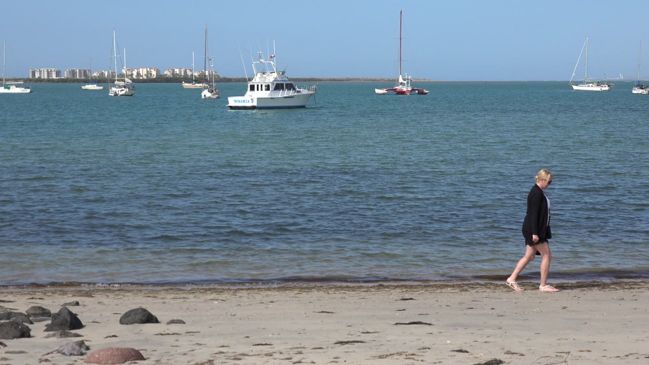 The woman has moved farther across the Los Cabos shoreline, with low waves behind her and a white boat, sailboats, and distant buildings along the horizon.