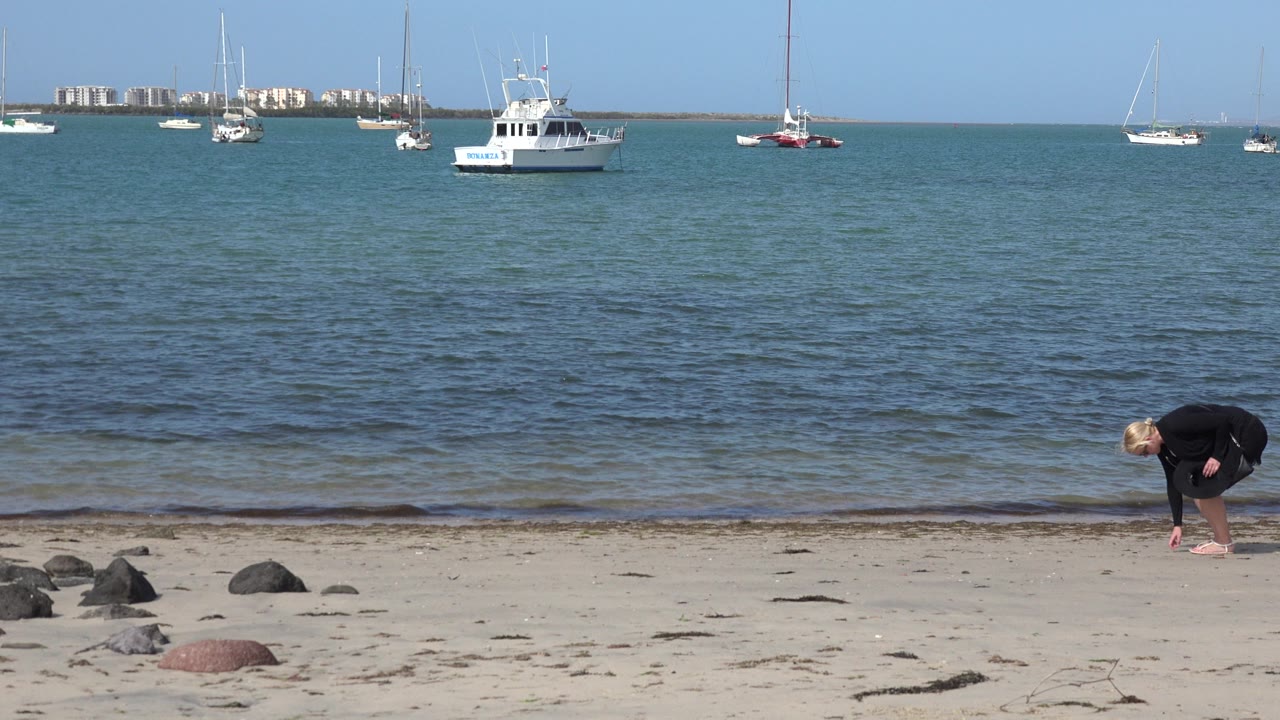 Woman bending on a sandy beach with boats offshore.
